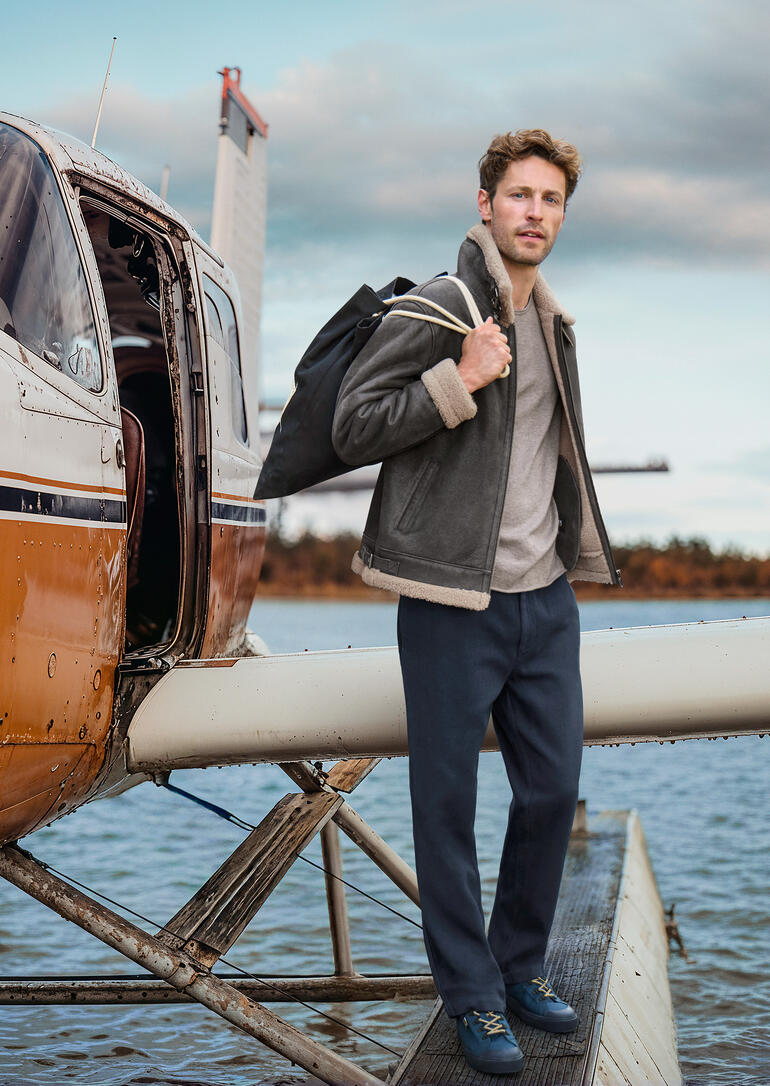 Man in bruine B3 schapenvacht pilotenjacke staat naast een vliegtuig.