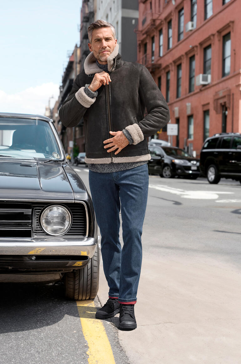 Man in schapenvacht pilotenjack poseert naast een klassieke auto