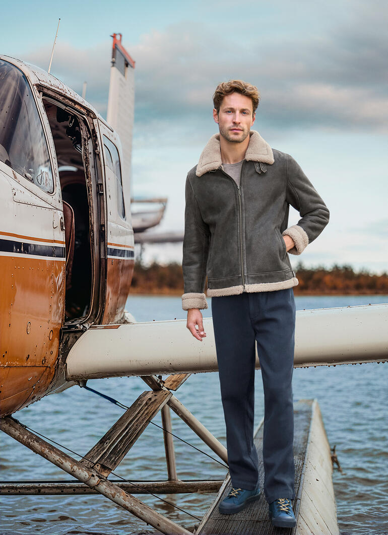 Man in bruine B3 schapenvacht pilotenjacke staat naast een vliegtuig.