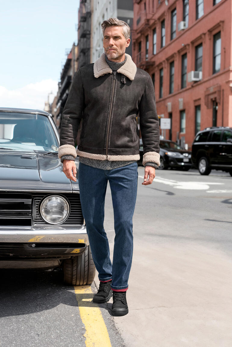 Man in een bruine lammy-pilotenjack staat voor een oldtimer in de stad.
