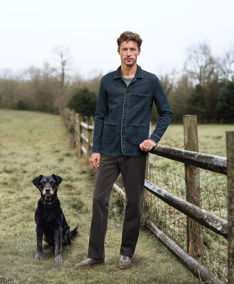 Man draagt een kasjmier denim shacket en staat naast een hond op een weide.