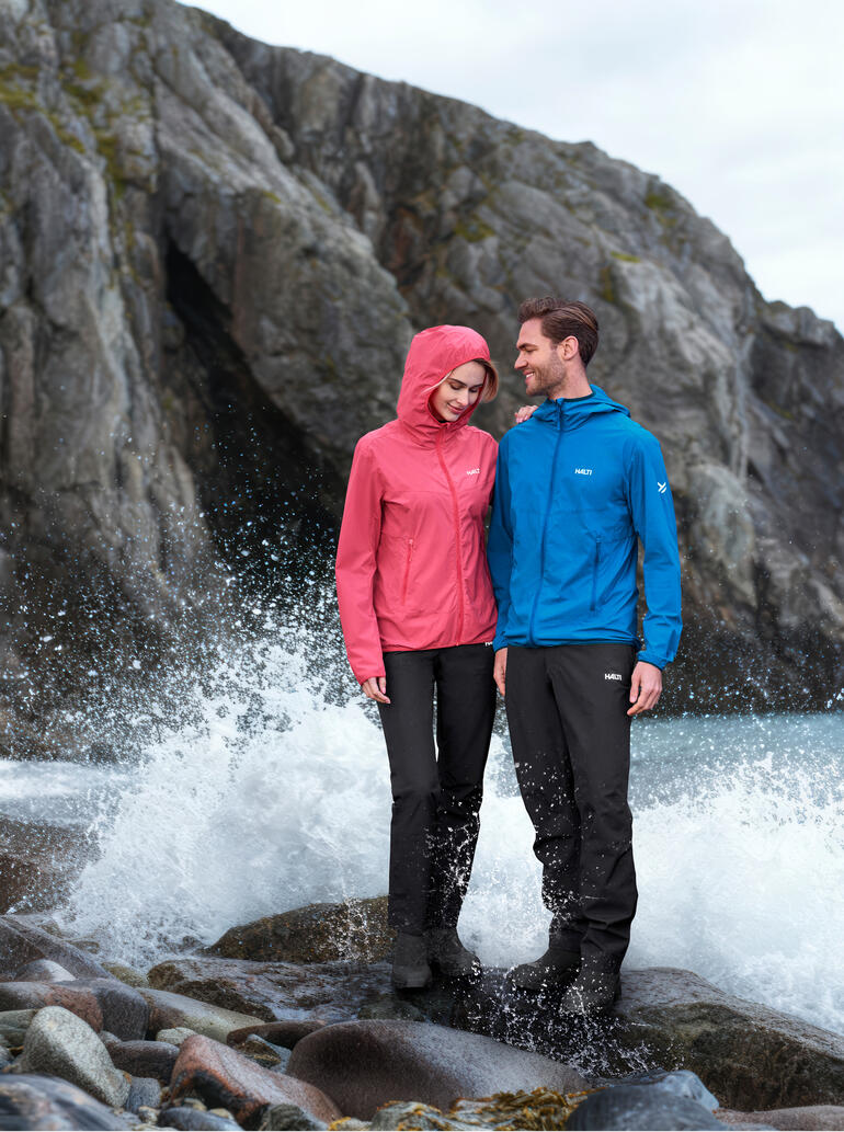 Koppel in outdoor kleding, een persoon draagt een roze softshell jack, de ander in een blauwe jack naast woelig water.