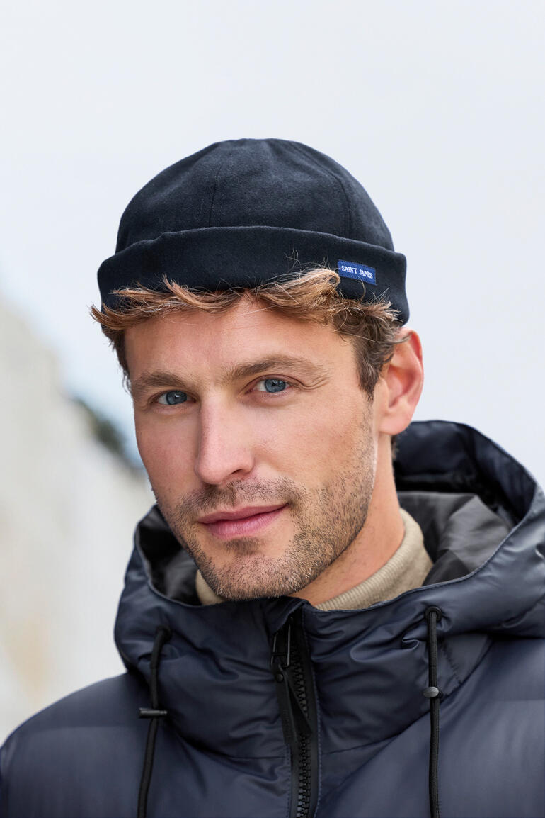 Man met een muts in een winterjas, glimlachend voor een onscherpe achtergrond.