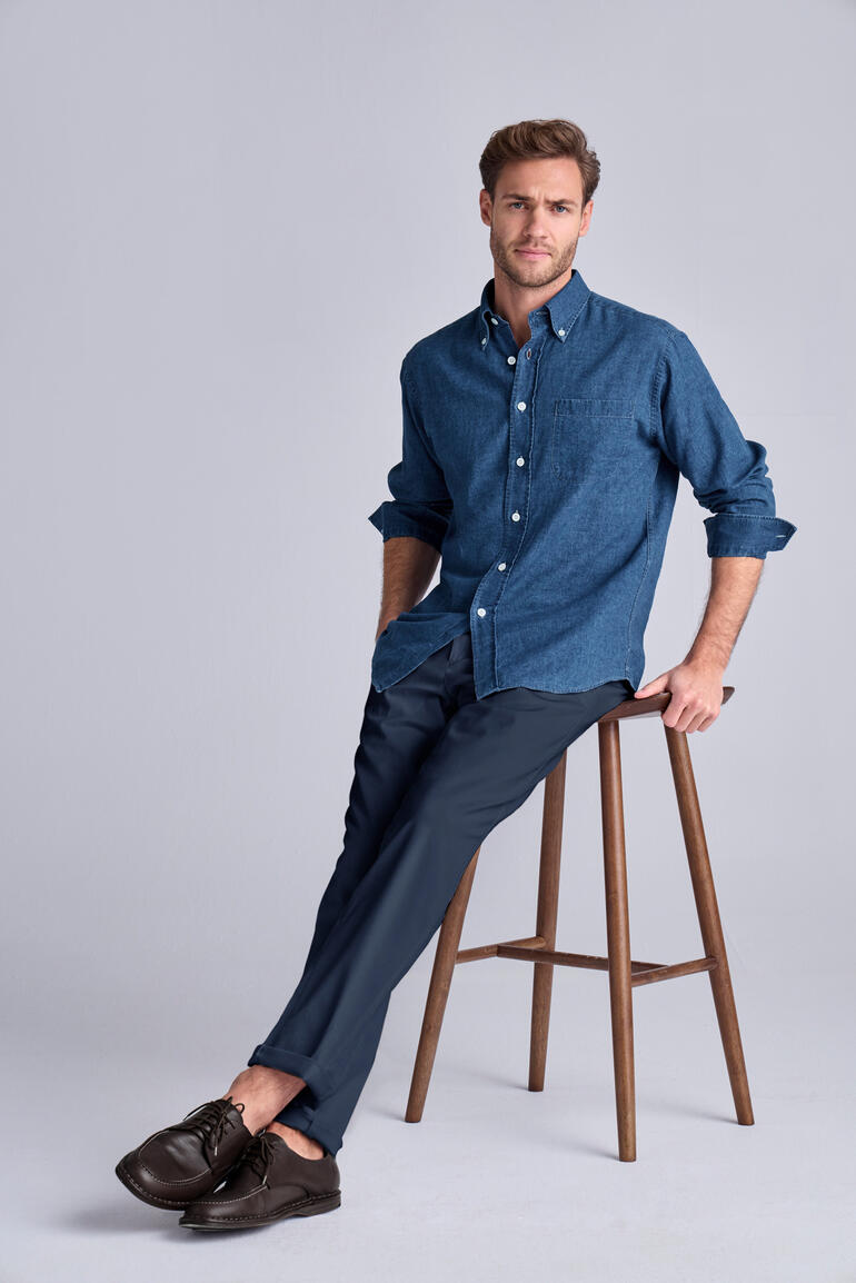 Man zit op een kruk en draagt een blauw shirt en een donkerblauwe chino broek.