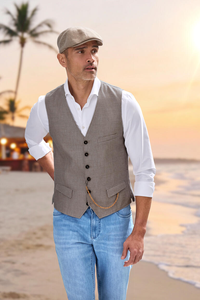 Man in een lichtbruine vest en wit shirt op het strand bij zonsondergang.