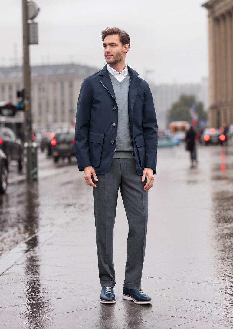 Donkerblauwe all-weather jas voor heren, staand op een regenachtige straat.