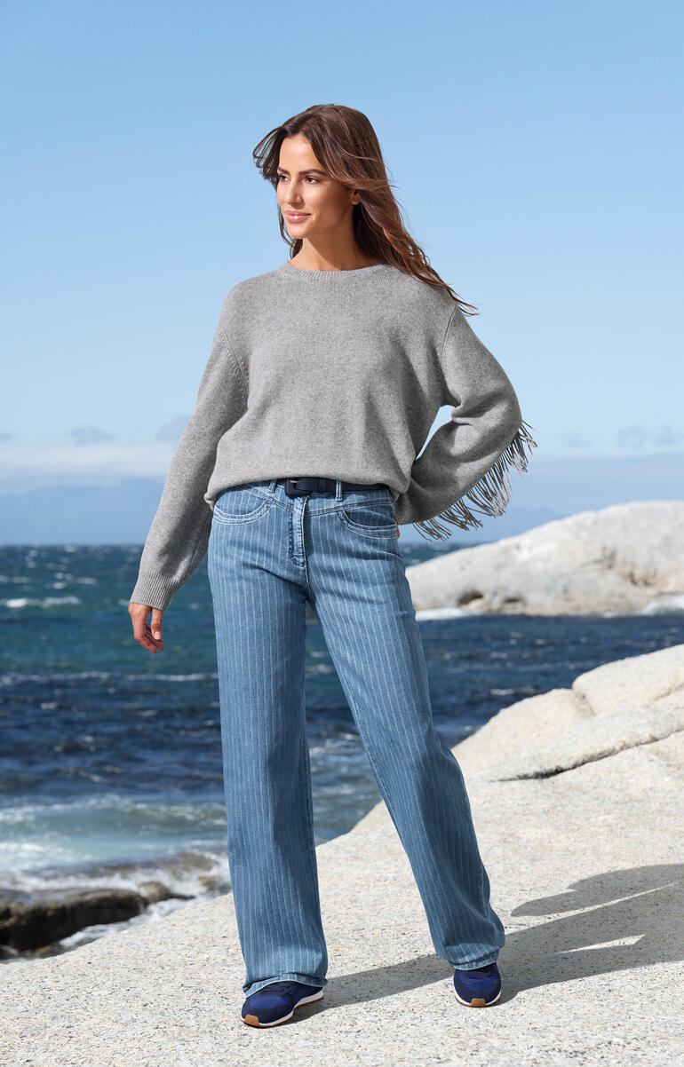 Grijze trui met franjes, casual over jeans aan het strand gedragen.