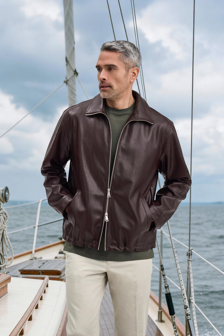 Man in bruine kunstlederen jack met grijze trui op het dek van een zeilboot