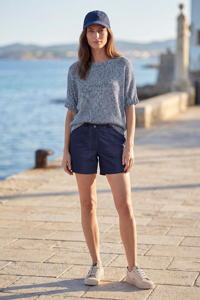 Vrouw staat aan de zee in een blauwe trui en shorts.