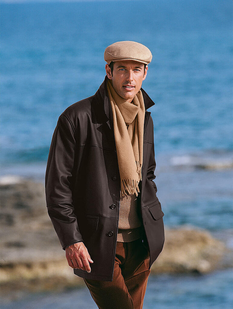 Man met een kameelhaar pet en sjaal met uitzicht op de zee.