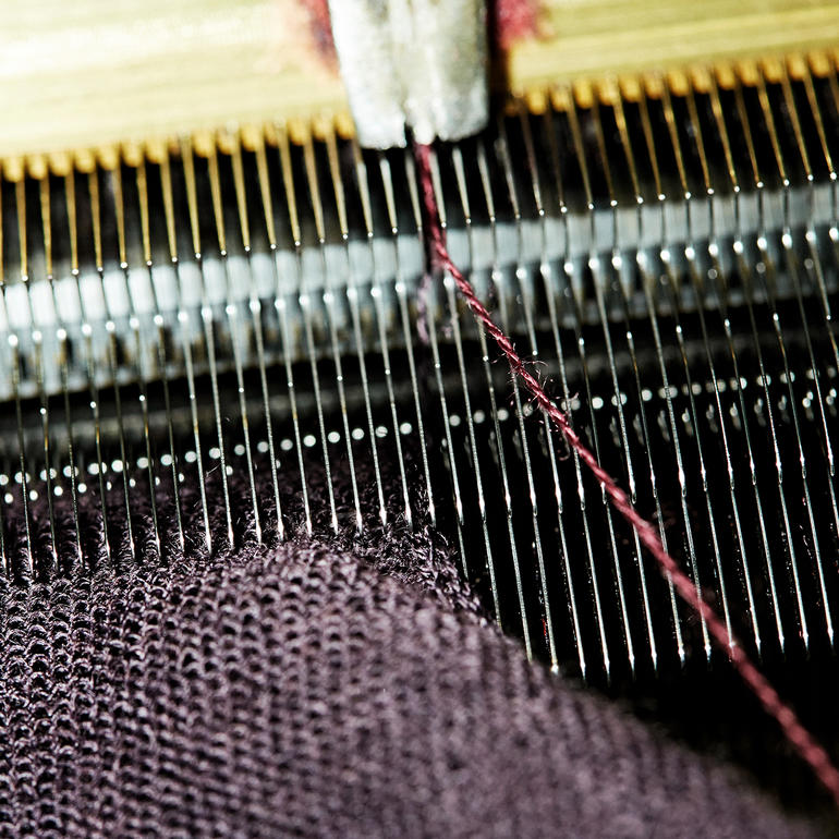 Slechts een handvol breimodeateliers in de wereld beheersen het gecompliceerde 30-gauge-breiproces: met 30 naalden per inch is het fijner dan standaard fijn breien (18-gauge). Detailopname van een breimachine met garen