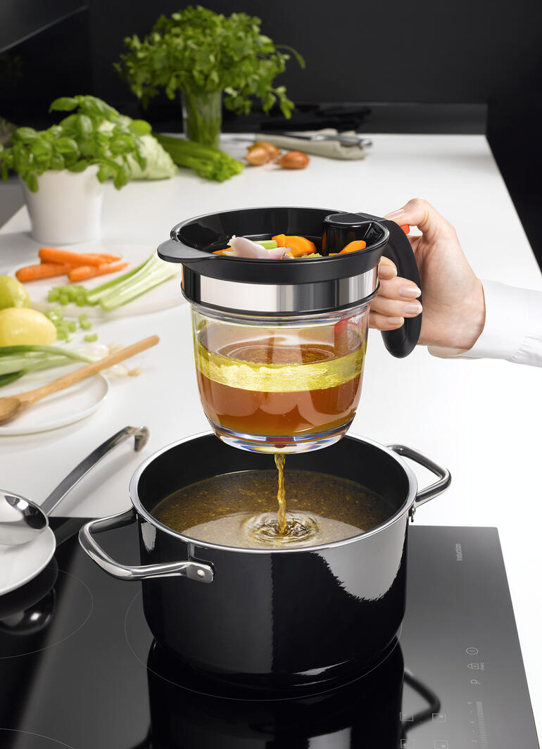 Hand met een zeef giet groentebouillon in een pan, terwijl een keuken op de achtergrond zichtbaar is.