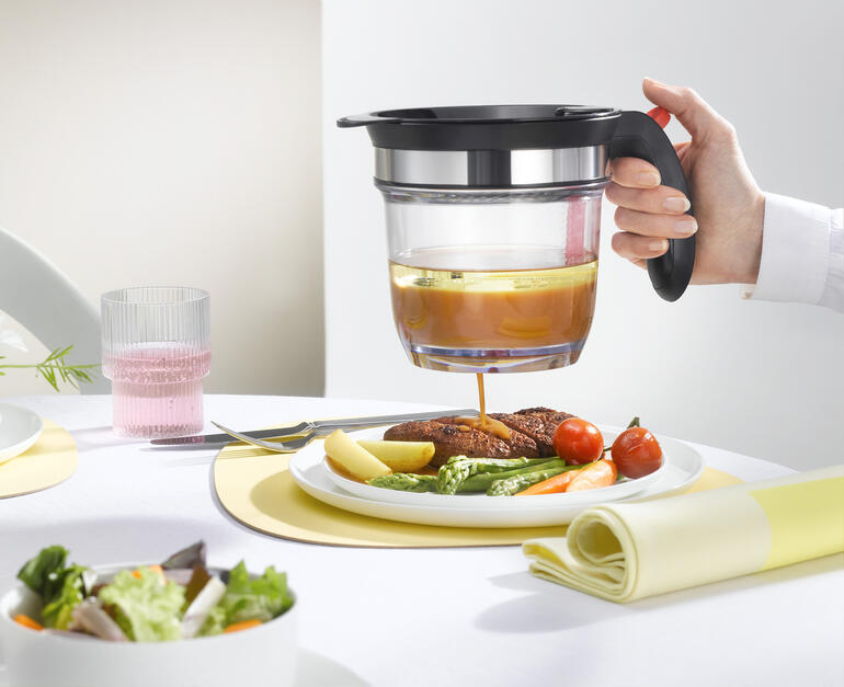 Hand met een zeef die bouillon over een bord met groenten en vlees giet, terwijl een gedekte tafel op de achtergrond staat.