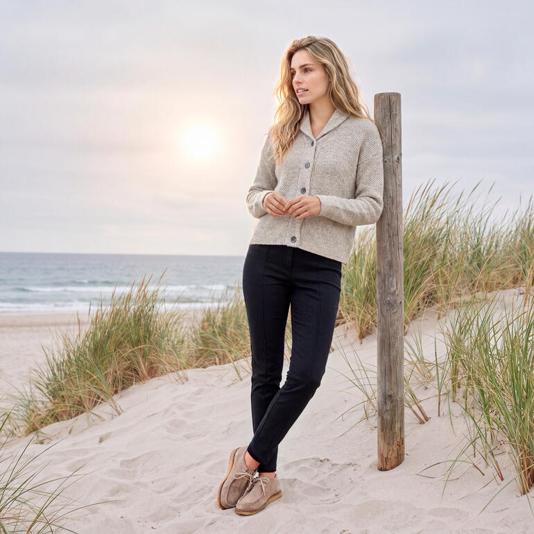 Vrouw in een grijze cardigan staat op het strand met zonsondergang op de achtergrond.